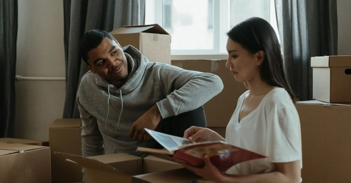 Un couple explore leur nouvelle maison tout en déballant des cartons et en consultant un album photo.