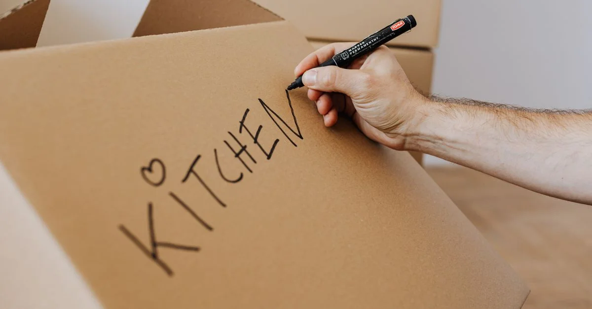 Une personne étiquetant une boîte en carton comme 'Cuisine' lors d'un déménagement.
